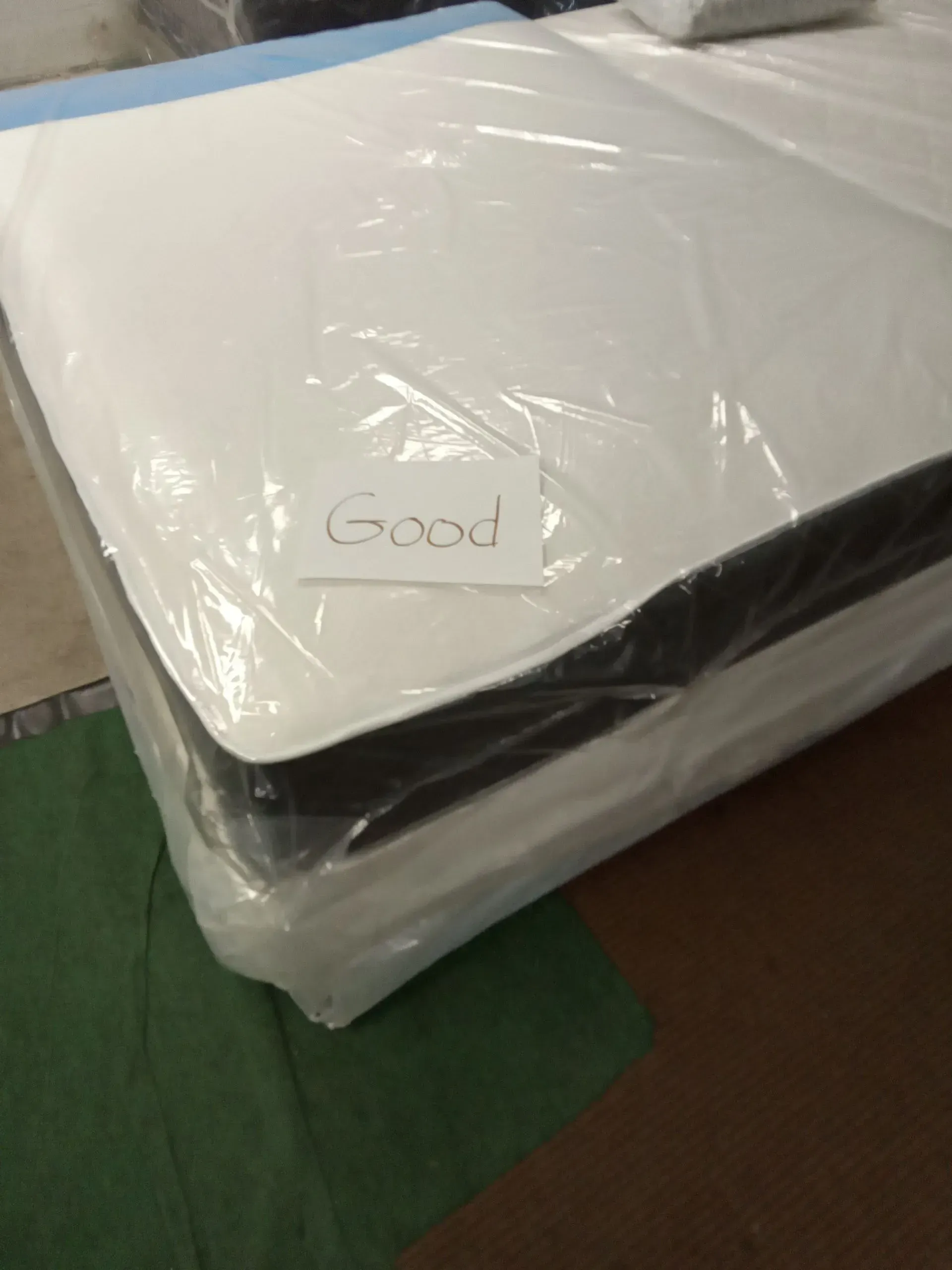 Plastic-wrapped white mattress or cushion with a small “Good” note on top