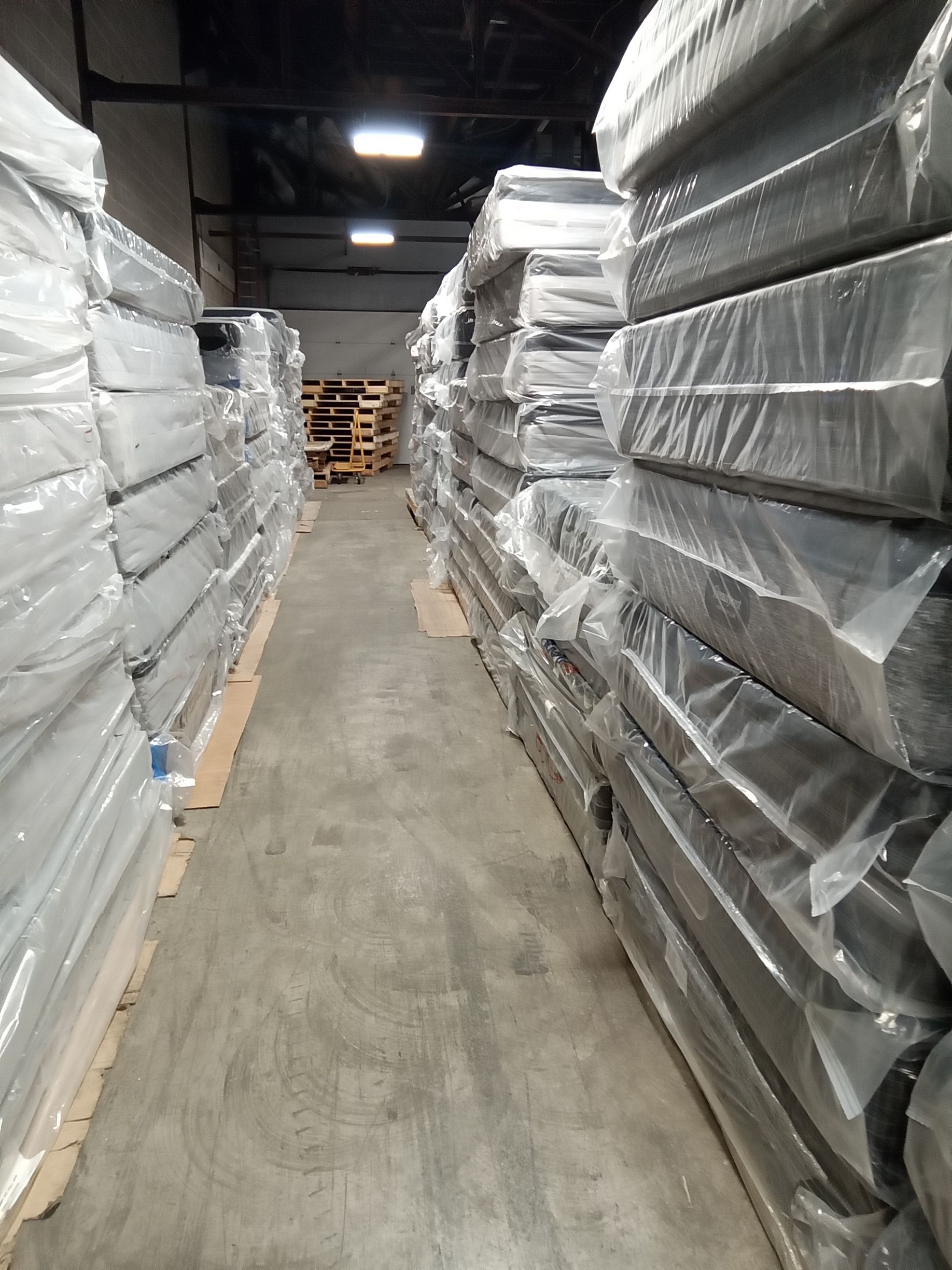 Narrow warehouse aisle lined with shrink-wrapped pallets on both sides and stacked pallets at the far end