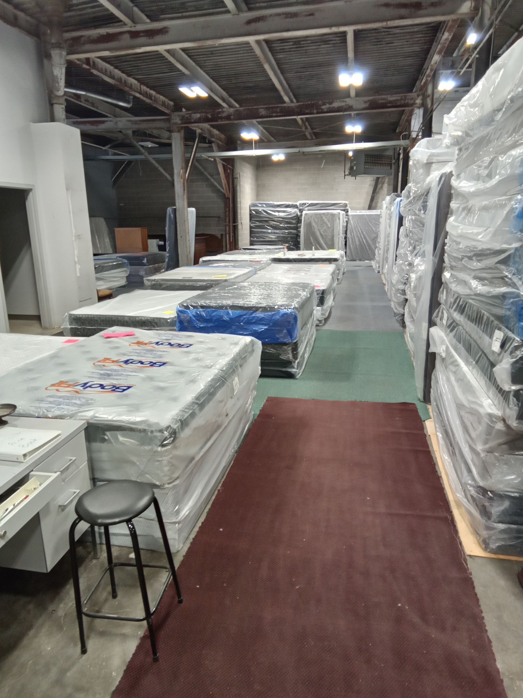Warehouse with stacked wrapped mattresses, a red carpet aisle, and a stool in the foreground.