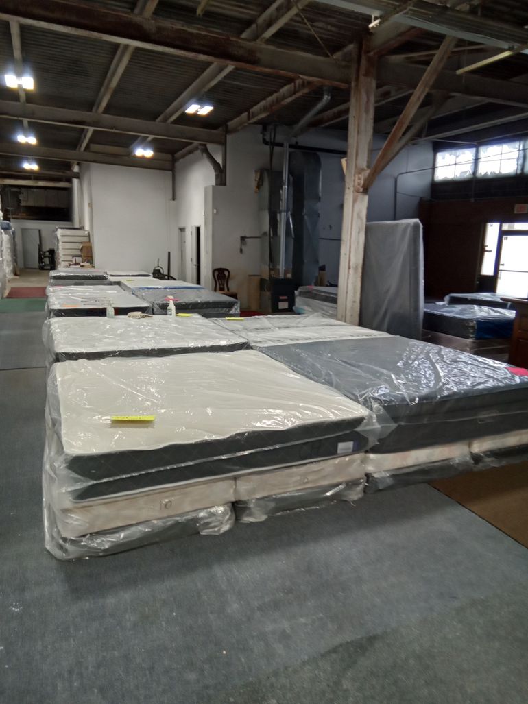 Warehouse with stacked plastic-wrapped mattresses or foam panels on pallets under exposed beams.