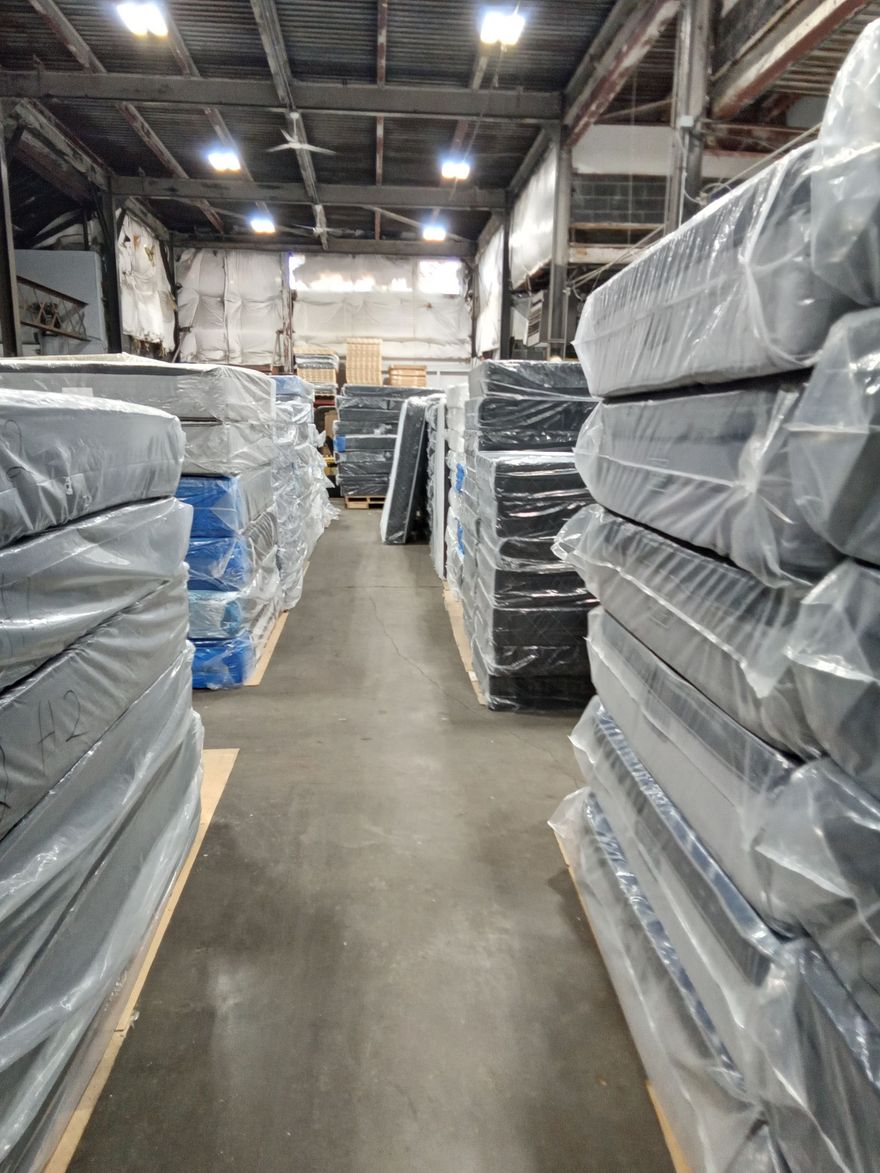 A warehouse aisle lined with stacked, plastic-wrapped mattresses or cushions on both sides.