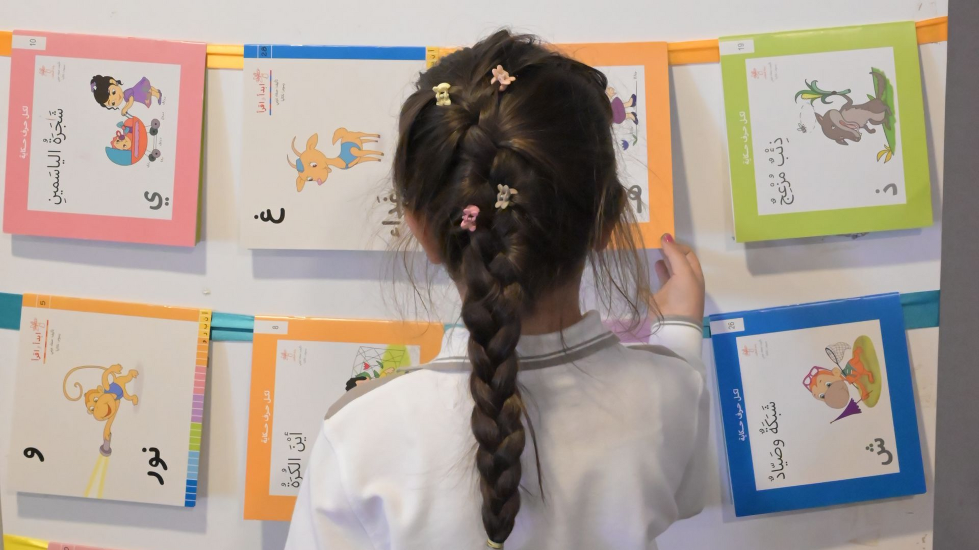 Child with braided hair stands facing a colorful wall display of drawings and schoolwork.