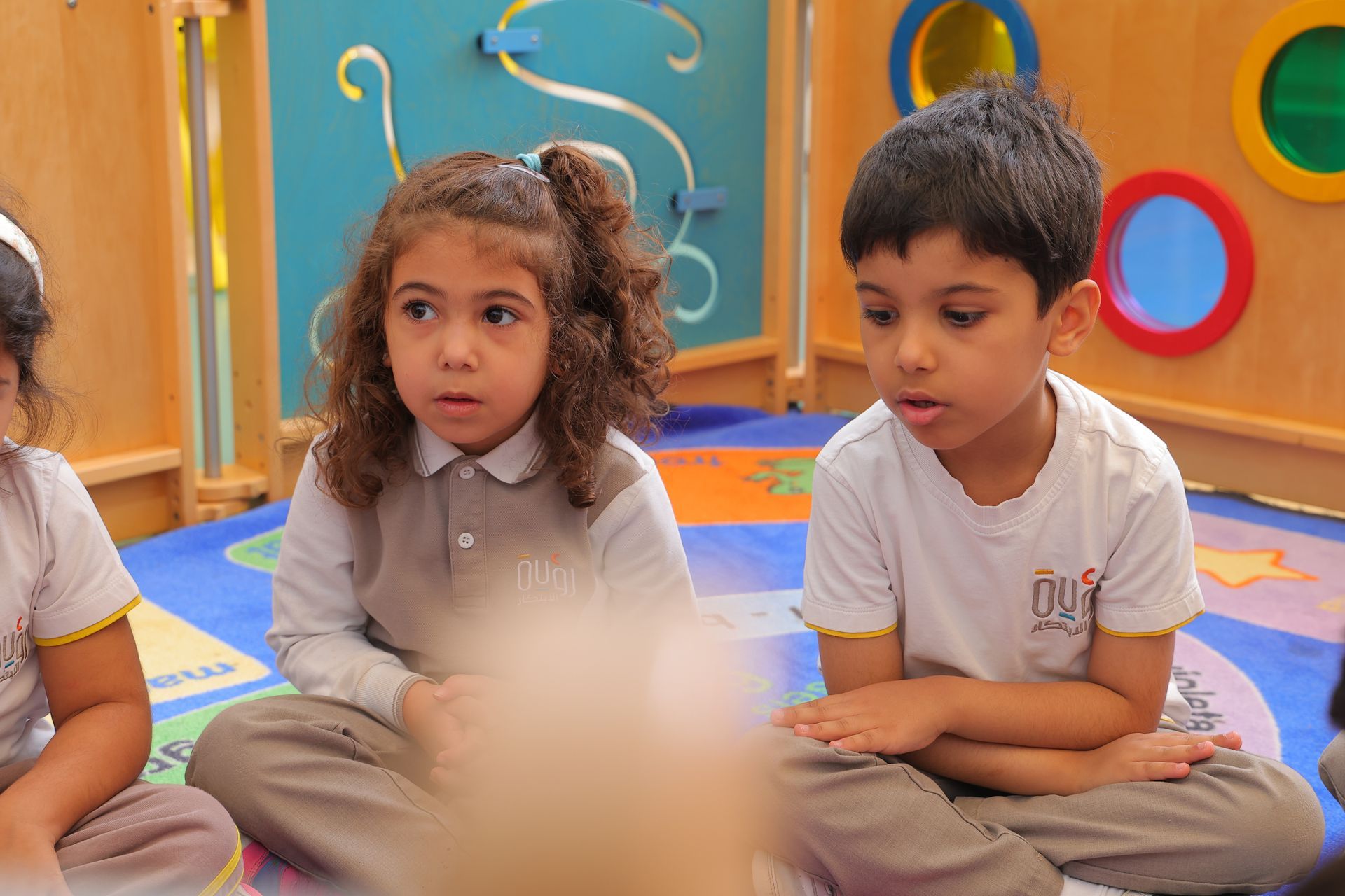 Two children sitting cross-legged on a colorful classroom floor, looking ahead attentively.