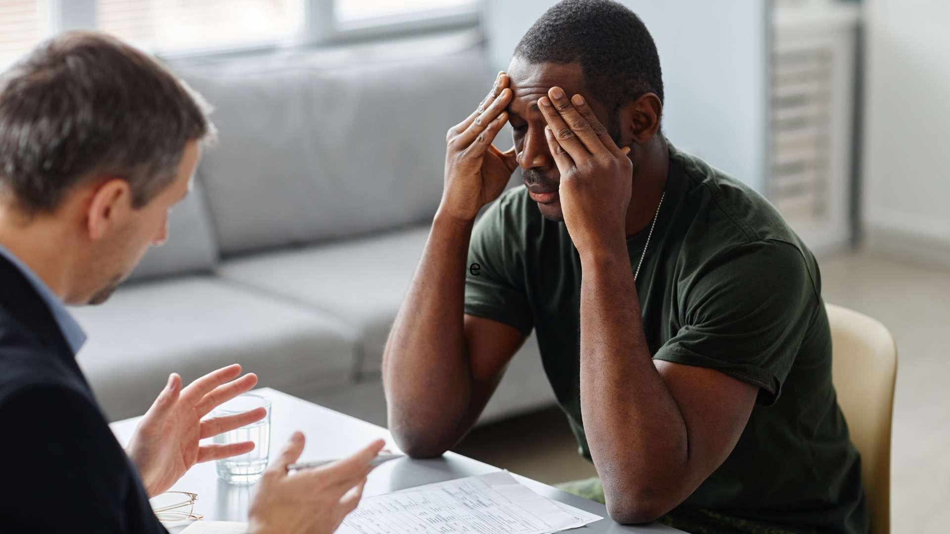 A man is sitting at a table with his head in his hands while talking to another man.