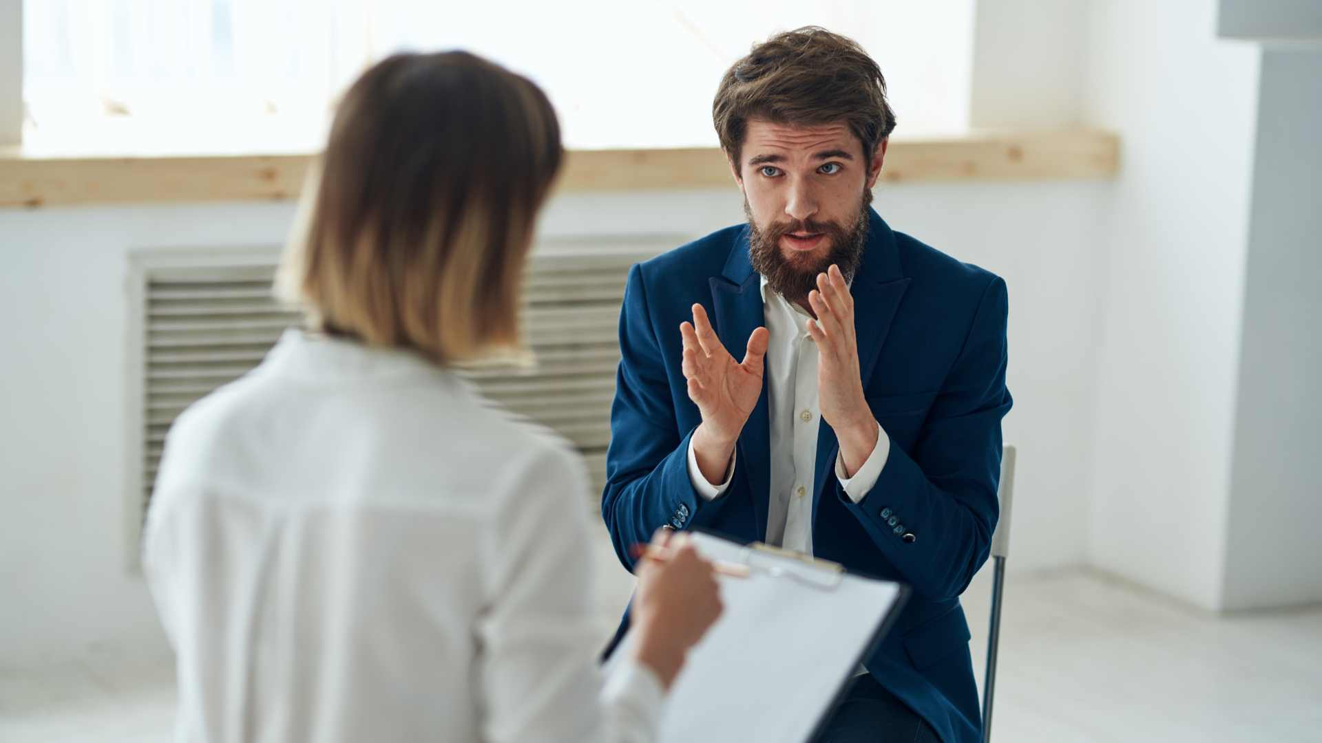 man holding both of his hands up as he speaks to a psychiatrist sitting on a chair across him