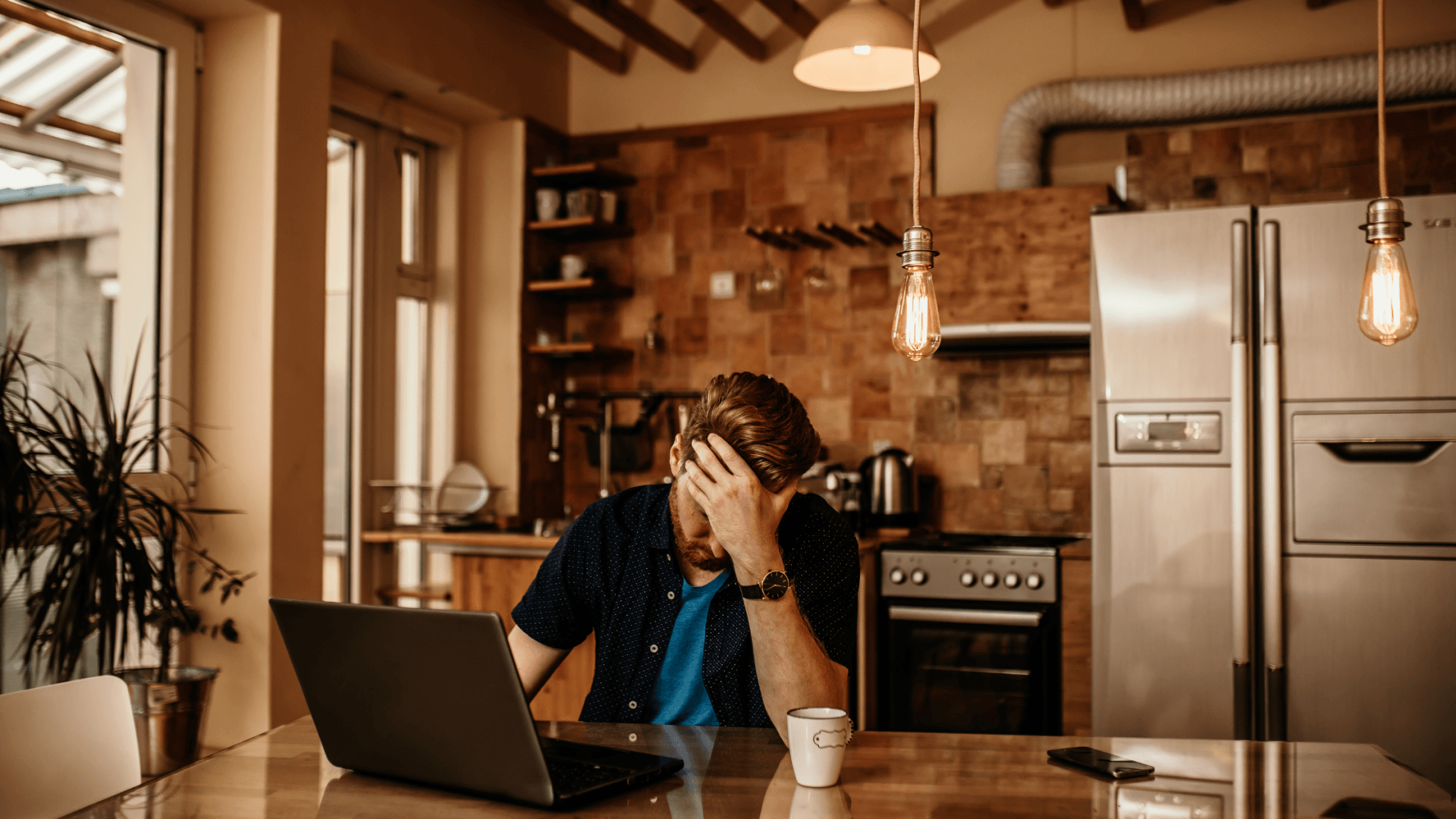 man with his palm on his head while on his computer in the kitchen