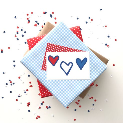 Een cadeaukaartje met daarop drie harten in rood, wit en blauw op een stapel cadeautjes