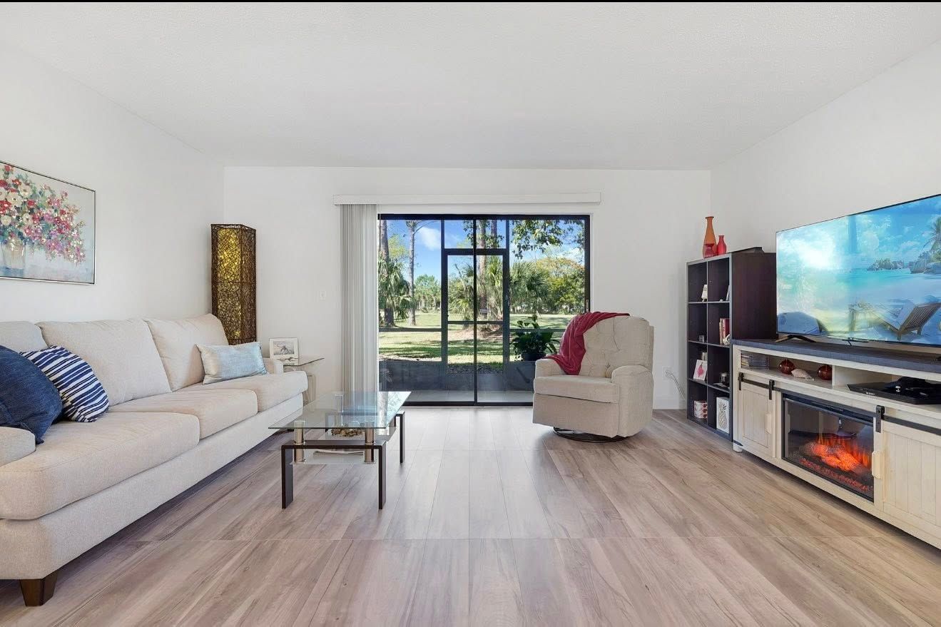 Living room with sofa, armchair, TV, and window overlooking a green area.