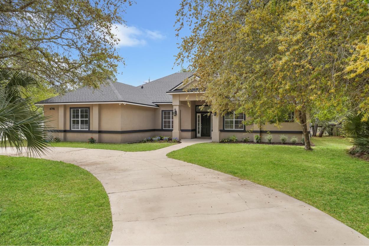 Single-story house with a curved driveway, green lawn, and large trees in front.