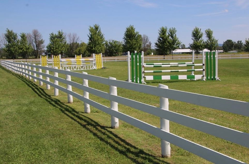 White fence lines a grassy horse arena with colored jump obstacles under a clear sky