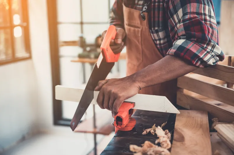 The Handyman Winnipeg using a circular saw to cut a wooden plank in a workshop; sawdust visible.