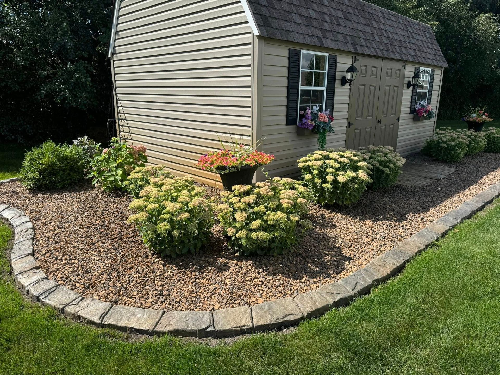 Small beige garden shed with flower beds, gravel border, and curved stone edging in a landscaped yard