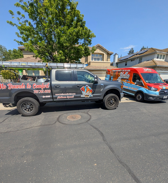 Two parked branded pickup trucks on a sunny street beside a tree and house.