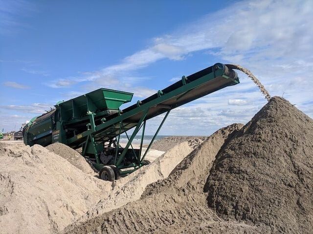 Une machine de convoyage industrielle verte dépose un gros tas de sable sur une plage sous un ciel bleu.