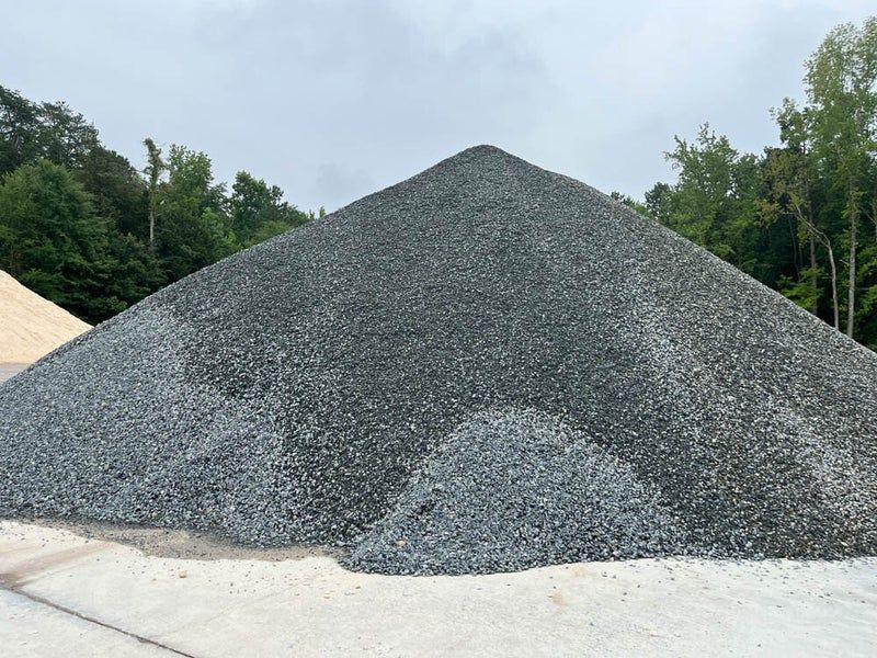 Un grand tas conique de pierres concassées grises repose sur une surface pavée, avec des arbres en arrière-plan sous un ciel nuageux.