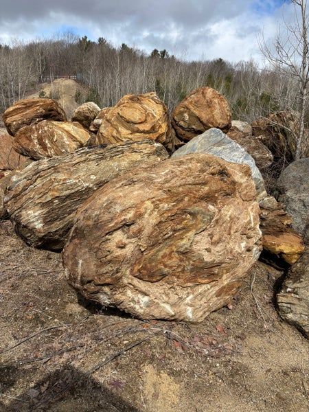 Un tas de gros rochers beiges et texturés repose sur la terre battue, au pied d'une colline boisée, sous un ciel nuageux.
