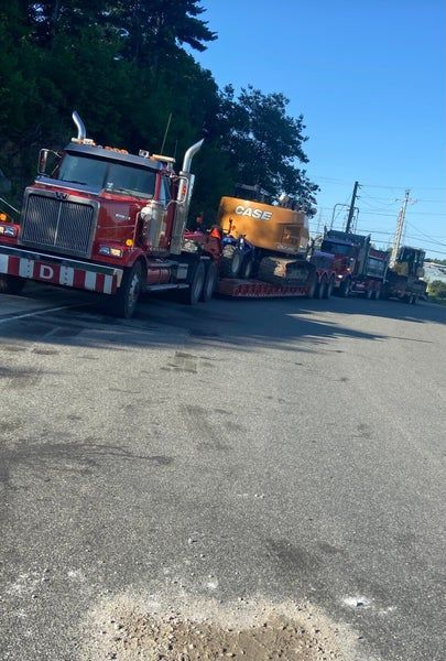 Un semi-remorque rouge tirant une remorque à plateau avec une pelle mécanique Case sur un terrain asphalté, suivi d'autres camions de chantier.