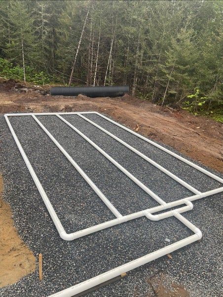 Grille de tuyauterie en PVC blanc disposée sur un lit de gravier dans une zone boisée, probablement pour l'installation d'un champ d'épandage septique.