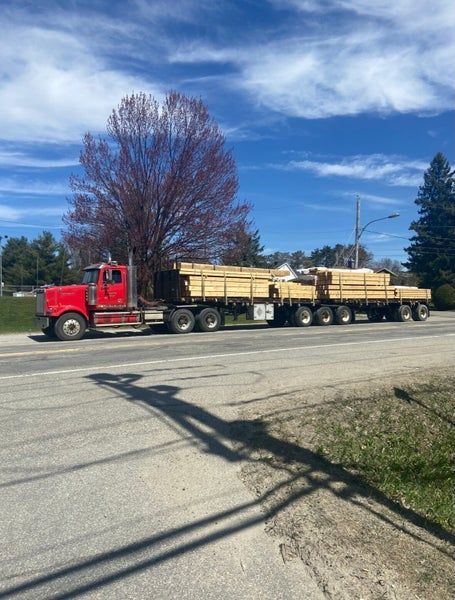 Un semi-remorque rouge tractant une longue remorque à plateau chargée de piles de bois sur une route ensoleillée.