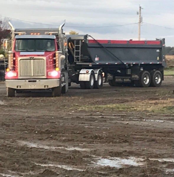 Un camion-benne rouge avec une remorque noire est stationné dans un champ boueux, en pleine campagne, par temps couvert.