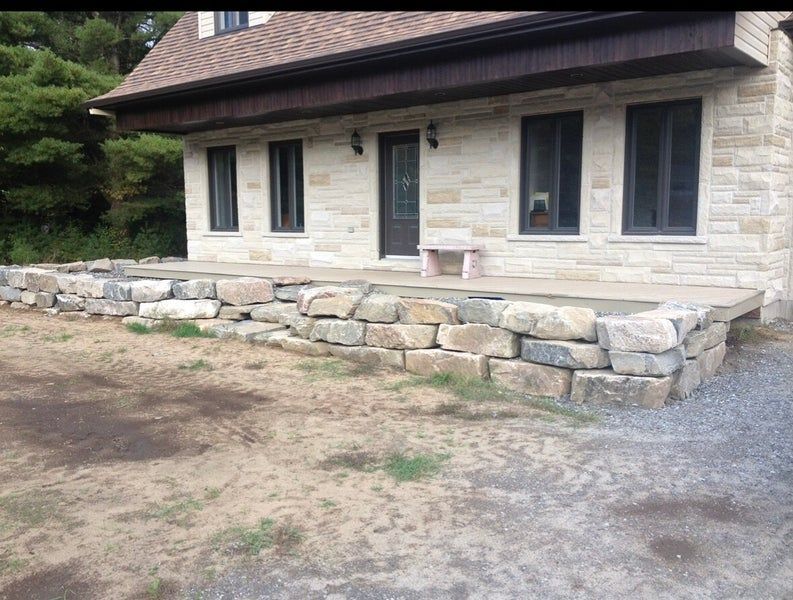 Une maison à façade en pierre, avec un grand mur de soutènement en roche naturelle et une terrasse en béton à l'entrée principale.