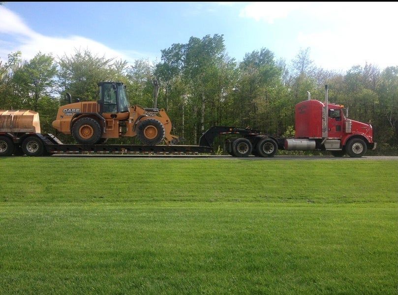 Un semi-remorque rouge transportant une chargeuse frontale John Deere jaune sur une remorque à plateau, stationné sur l'herbe contre une rangée d'arbres.