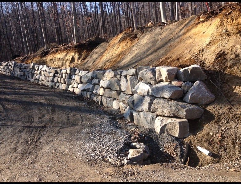 Un mur de soutènement en pierre en construction au pied d'un talus de terre dans une zone boisée.
