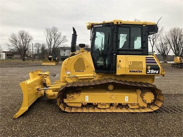 Un bulldozer Komatsu D39PX jaune, stationné sur un terrain gravillonné sous un ciel couvert.