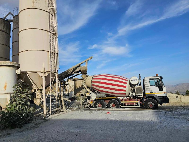 Un'autobetoniera accanto ai silos di cemento in un sito industriale sotto un cielo azzurro.