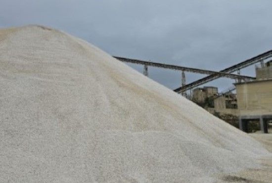 Un grande cumulo di ghiaia chiara in un sito industriale sotto un cielo nuvoloso, con nastri trasportatori ed edifici sullo sfondo.