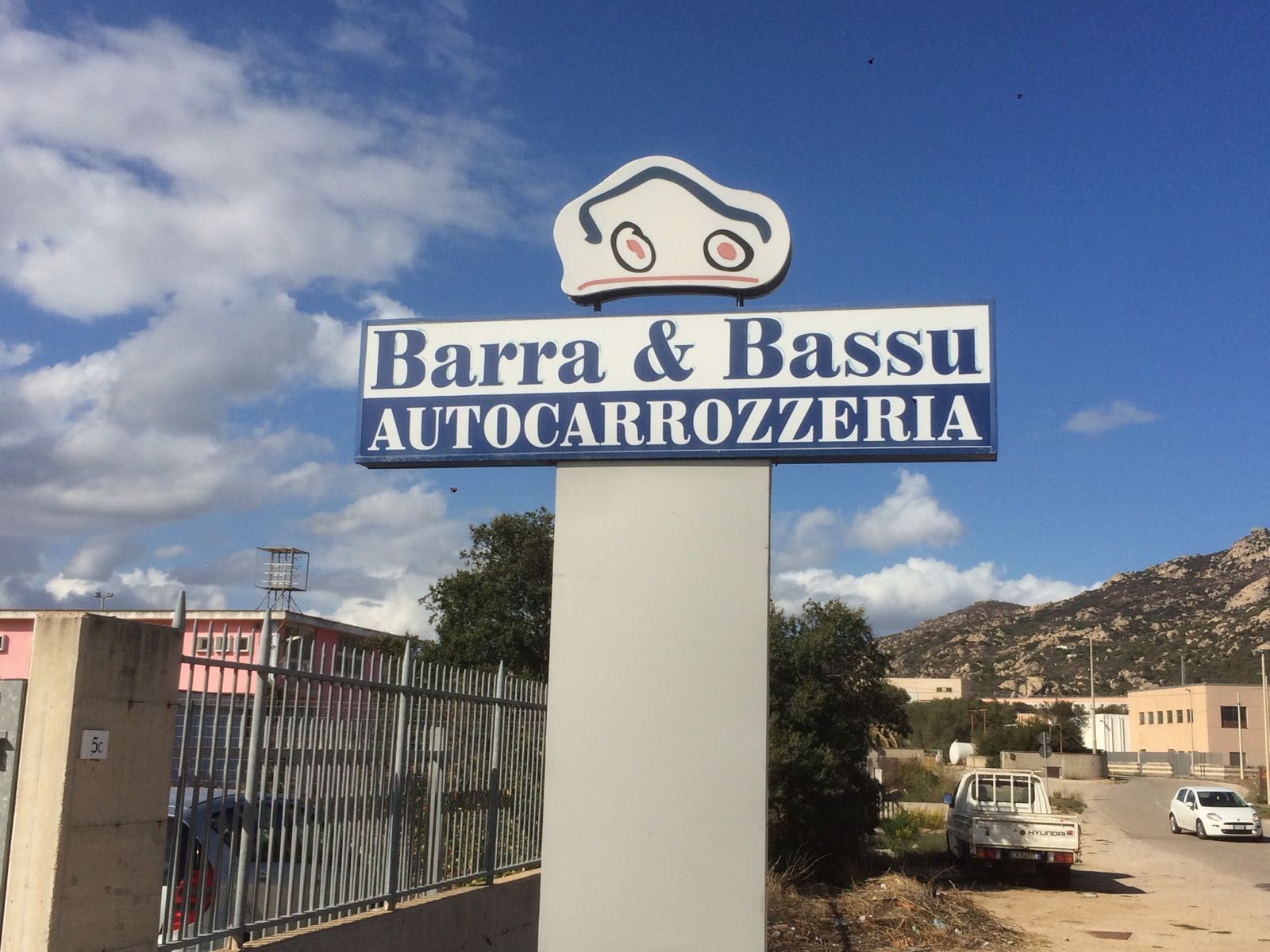 Insegna Barra & Bassu Autocarrozzeria su un pilastro bianco all'esterno di un'officina di carrozzeria sotto un cielo azzurro.