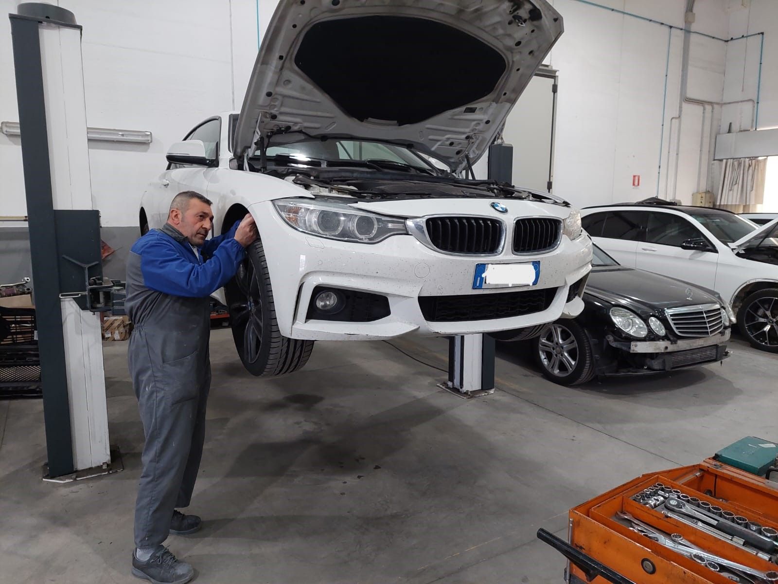 Un meccanico ispeziona una BMW bianca con il cofano aperto in un'officina, con un'altra auto nelle vicinanze.