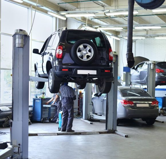 Un meccanico si trova sotto un SUV nero su un ponte sollevatore in un'officina, con un'altra auto nelle vicinanze.