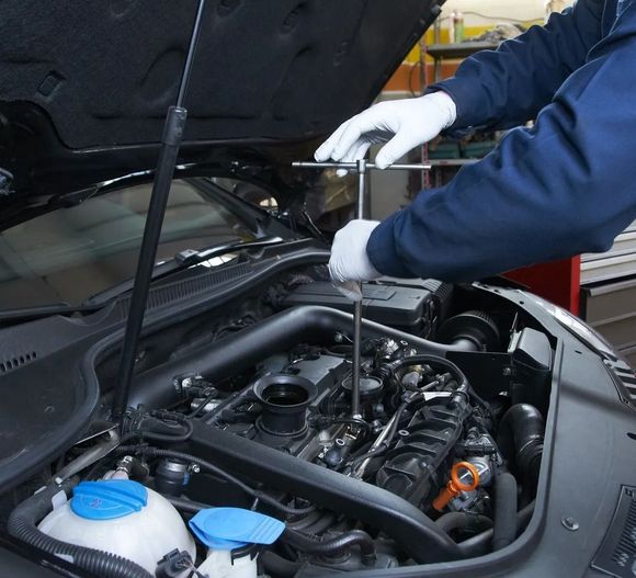 Meccanico al lavoro sotto il cofano di un'auto, nel vano motore, con attrezzi e tappi blu per i fluidi.