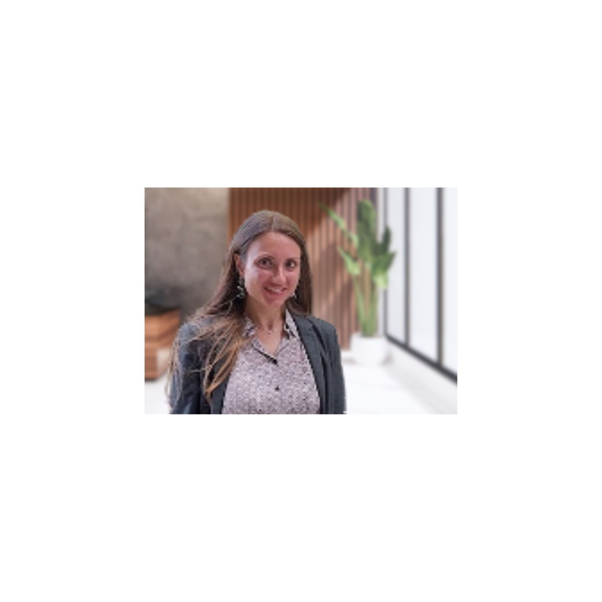 Woman in blazer and printed shirt smiles, standing near a window with plants.