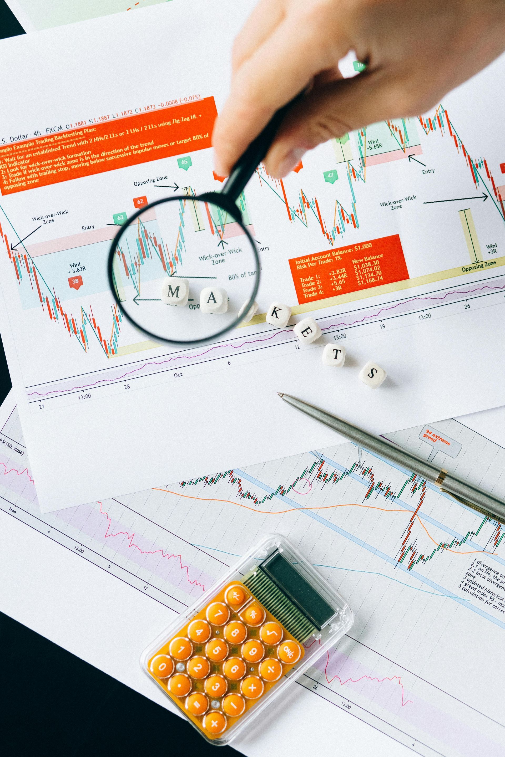 Hand using magnifying glass on financial charts with calculator and dice.