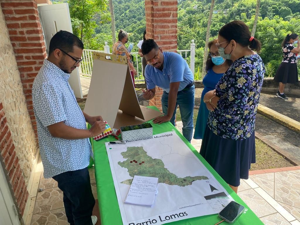 La gente se reúne alrededor de un mapa sobre una mesa al aire libre. Un hombre escribe, otros señalan y comentan; mesa verde, pared de ladrillos, árboles al fondo.