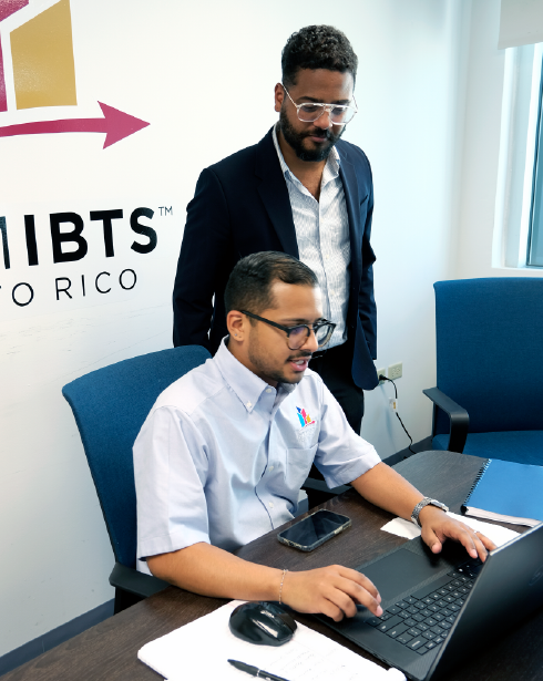 Two men are looking at a laptop in front of a wall that says iibts