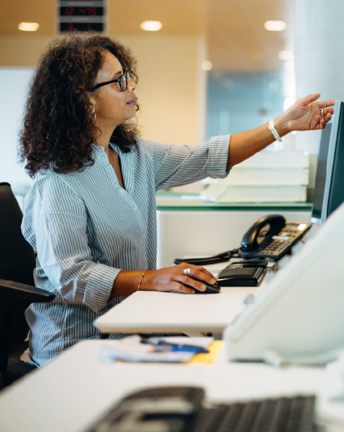 Una mujer con gafas, sentada en un escritorio, señala la pantalla de una computadora. Está en una oficina, con un teléfono y un teclado visibles.