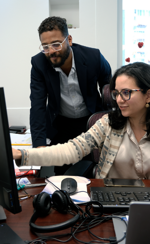 Un hombre con chaqueta y gafas se inclina sobre una mujer en un escritorio, ambos mirando una pantalla de computadora en una oficina.