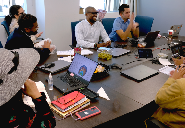 Personas sentadas alrededor de una mesa de conferencias, probablemente en una reunión de negocios. Un hombre negro con camisa blanca sonríe. Hay computadoras portátiles y otros artículos sobre la mesa.
