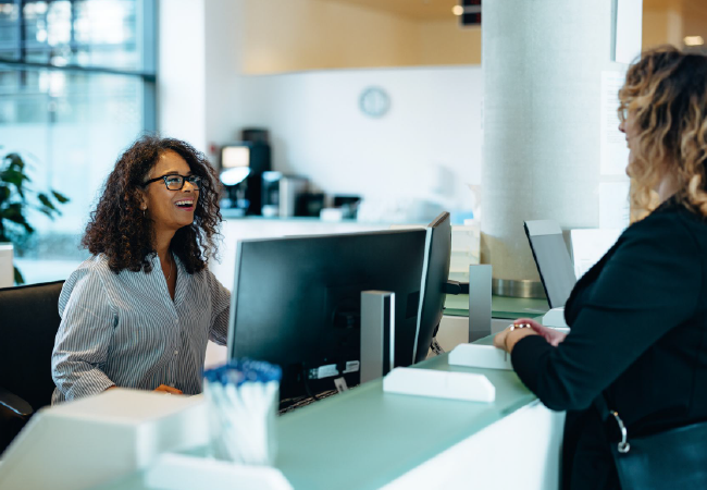 A woman is sitting at a desk in front of a computer talking to another woman.