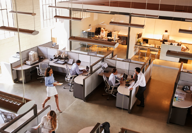 Vista aérea de una oficina moderna con empleados en cubículos, trabajando con computadoras. La gente camina y conversa en el espacio de trabajo abierto.