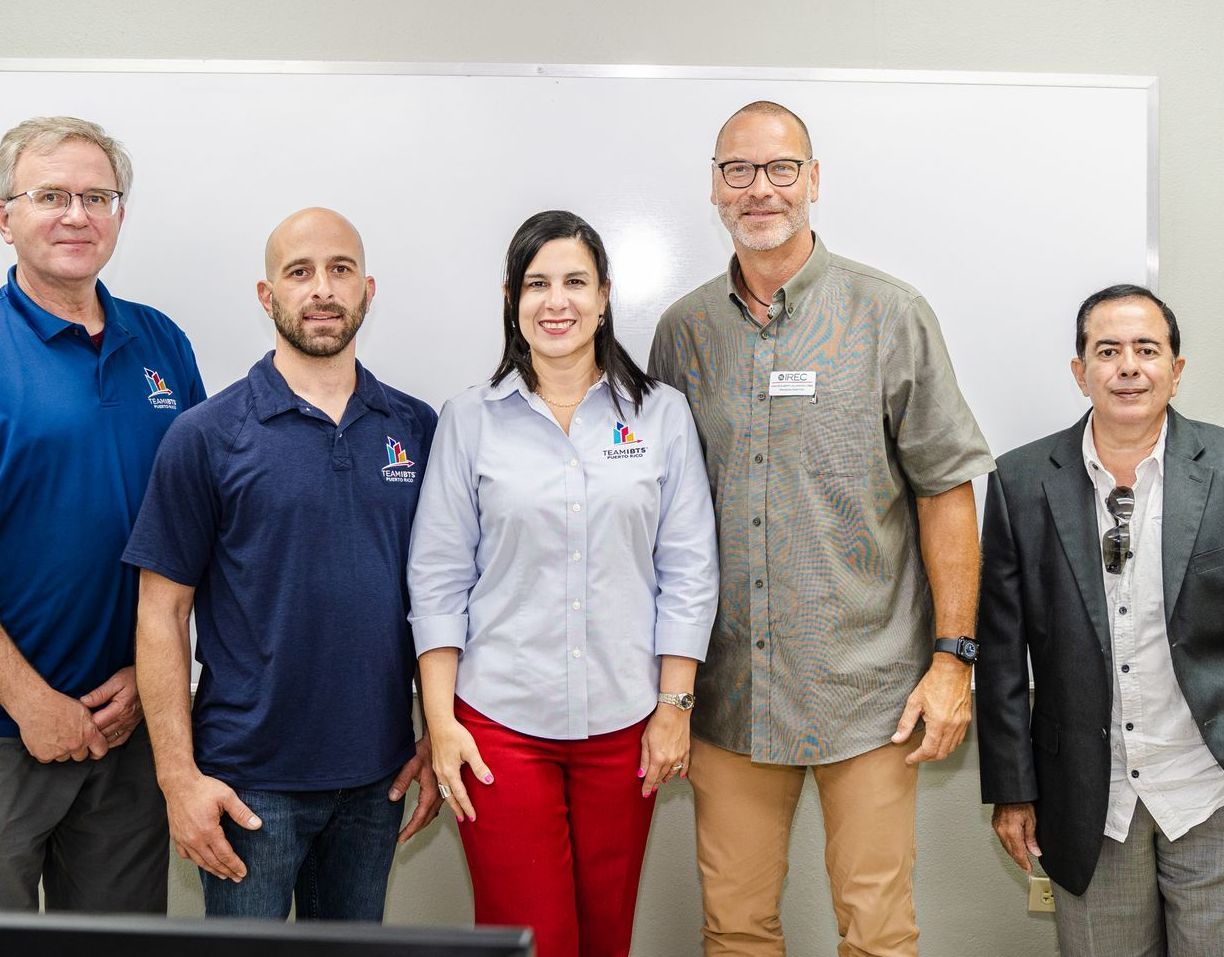 Cinco personas están de pie frente a una pizarra. Son diversas y visten ropa informal; la mujer del centro lleva pantalones rojos y una camisa azul claro.