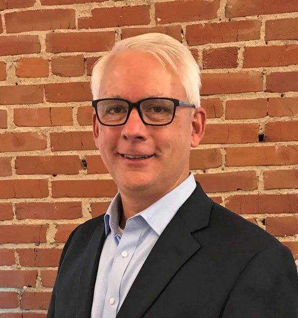 A man wearing glasses and a suit is standing in front of a brick wall.