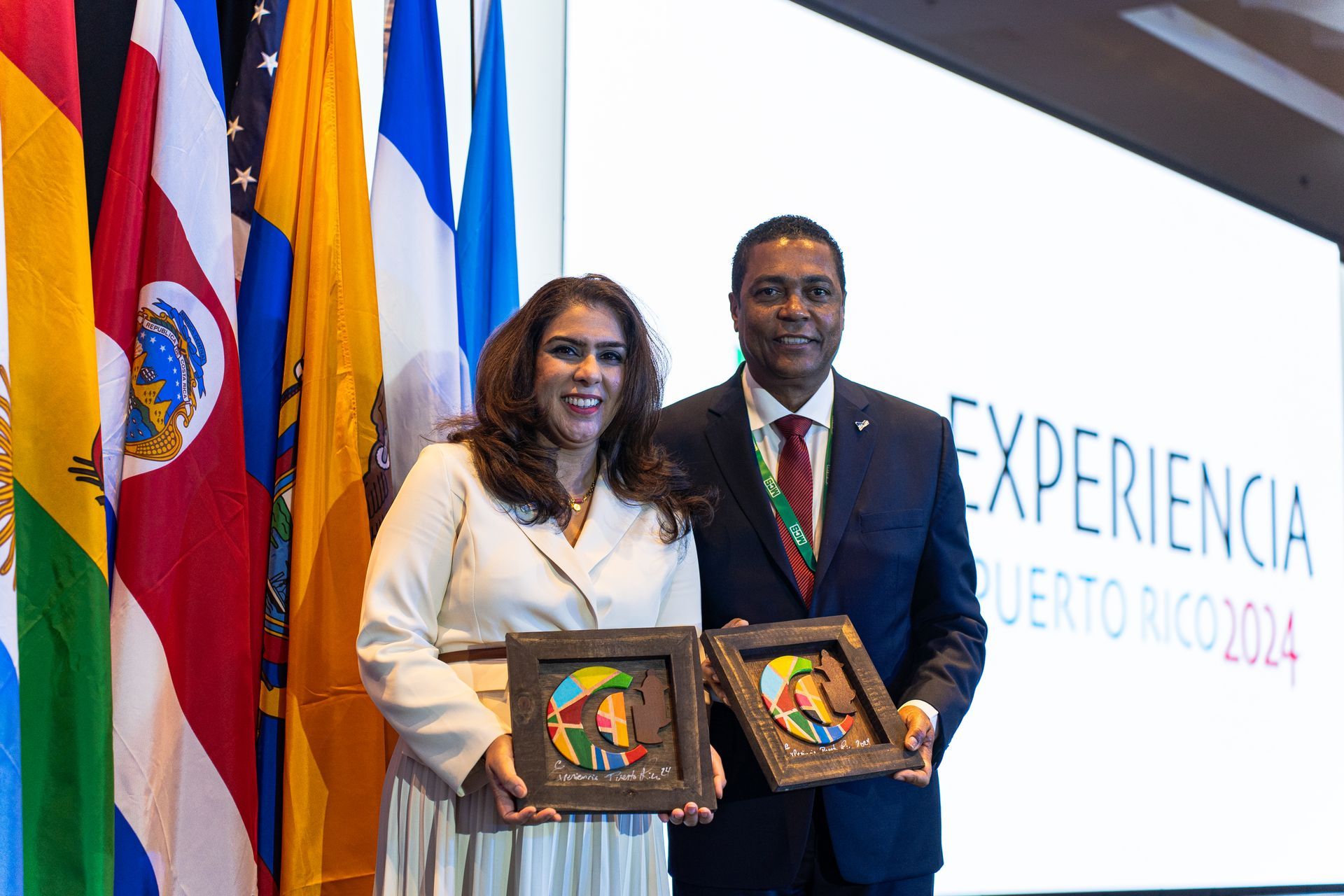 A man and a woman are holding framed pictures in front of flags.