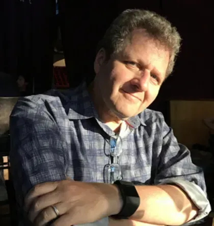 A man wearing glasses and a watch is sitting with his arms crossed and smiling.