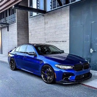 A metallic blue BMW M5 sedan parked on an asphalt street next to a light gray brick building.