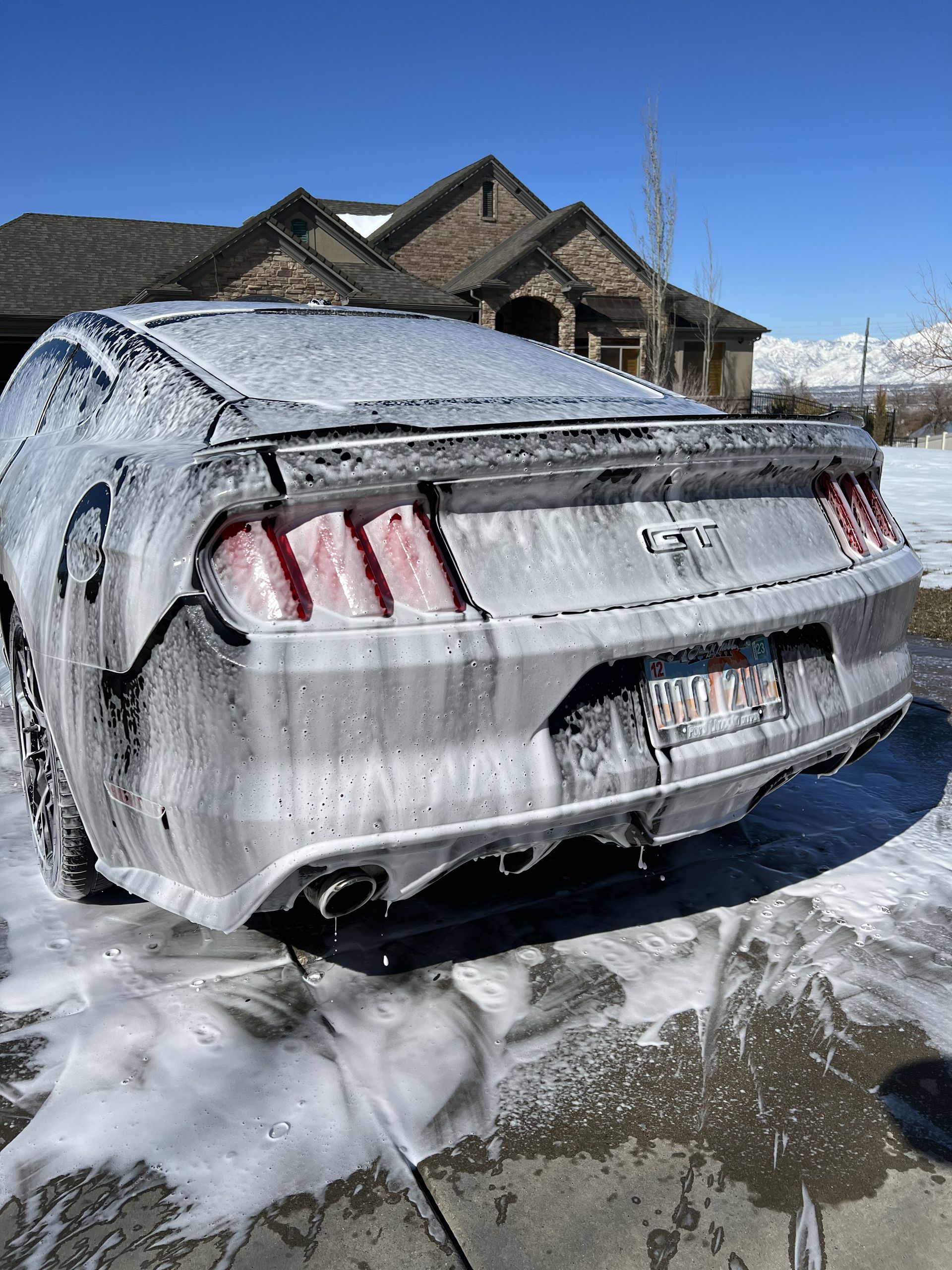 Silver Ford Mustang covered in car wash foam, outdoors near a house.