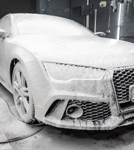 White Audi car covered in thick white foam, being washed at a car wash.